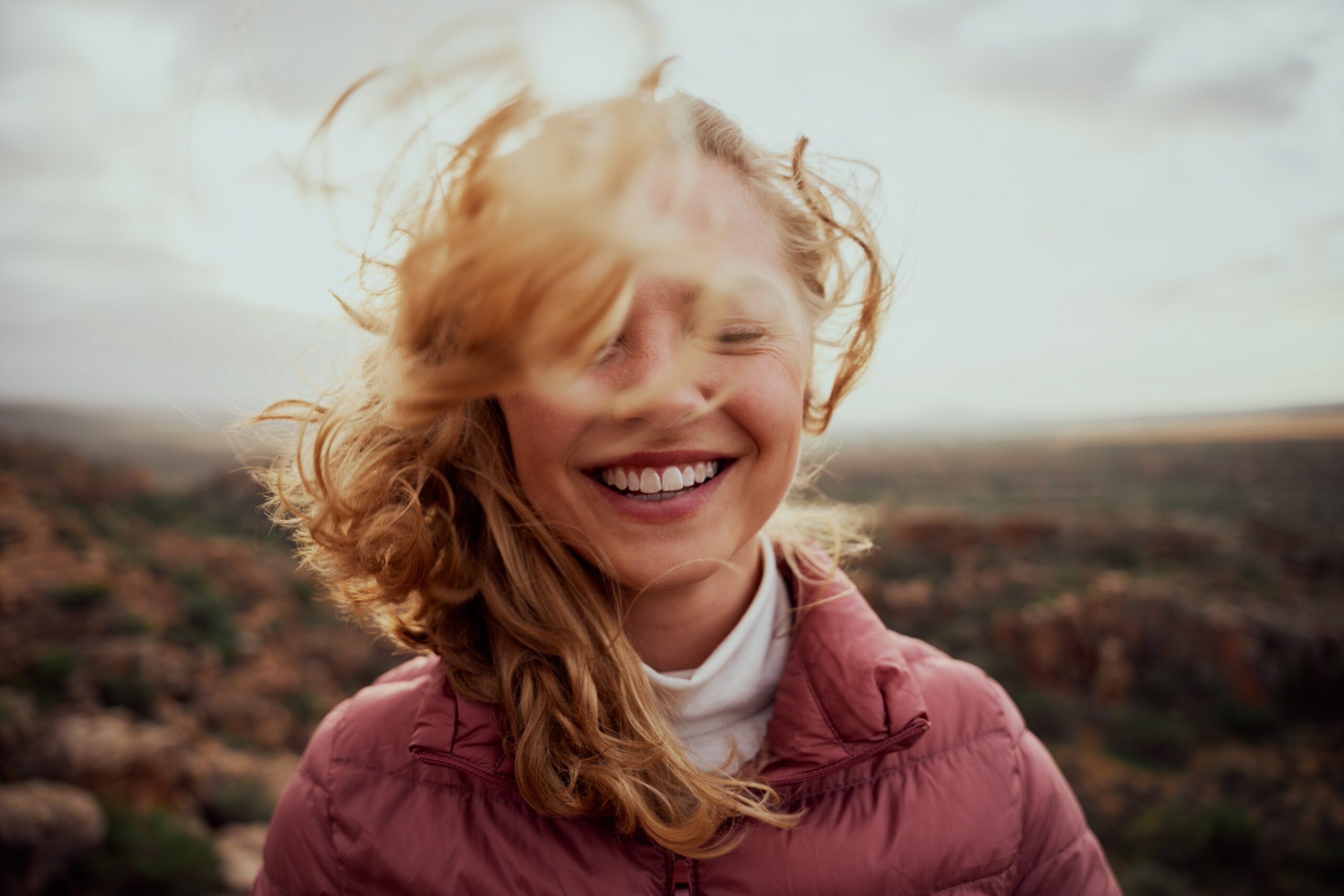 portrait-of-young-smiling-woman-face-partially-covered-with-flying-hair-in-windy-day-standing-at-mountain-carefree-woman